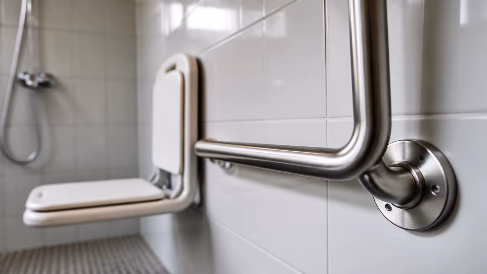 Détail d'une barre d'appui inox brossé scellée mécaniquement dans un mur carrelé blanc, avec siège de douche rabattable en arrière-plan